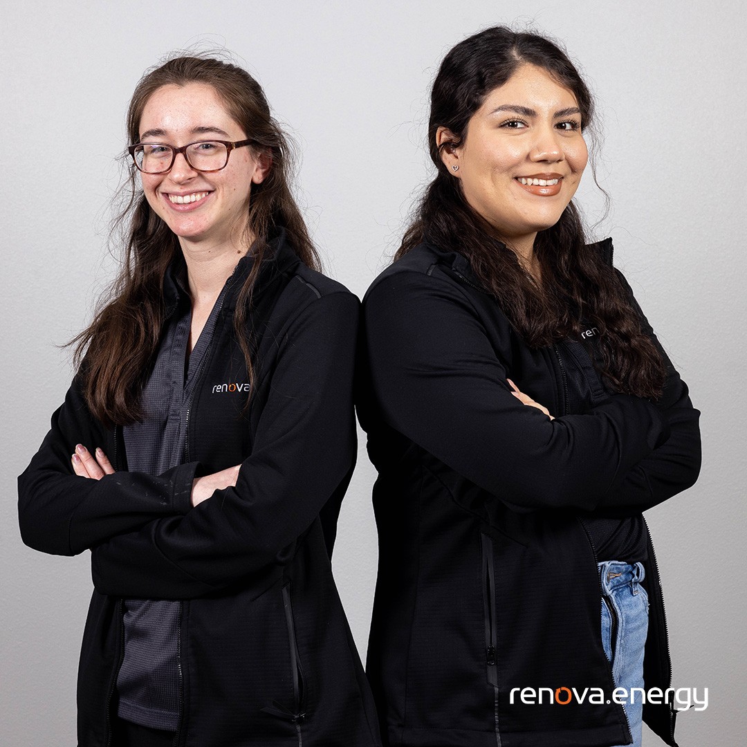 Photo showing 2 Smiling Renova Energy Female Employees. The employee on the left is crossing her arms smiling and is wearing prescription glasses and wearing a dark shirt under a long black long sleeve Renova jacket 