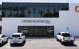 White building which has 2 tall palm trees on the left of the building with dark glass in front of second story. On top Renova Energy sign right below it with the o in Renova in the color orange and the rest of the sign in grey. There are palm trees showing in the glass reflection. There is a large orange O is on the very top right corner of the building on the first floor front doorway is all dark reflective glass. One white vehicle along with another white suv vehicle to the right of it totalling 2 vehicles is parked in front of building on the left then two empty parking spaces next to it on the right the next vehicle after the two empty parking spots to the right is a white pick up truck.