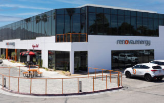 Outside of a white two story Renova Office Building with dark glass windows on the second floor. The front of the building has an orange metal rail alongside the concrete walkway area. There is an orange table with benches in front as well as another table with a black and red umbrella in front of the building where a Deli is located within the Renova building. There are 4 white vehicles parked out front with one that has the Renova logo shown on the side of the car. Renova.energy sign is on the building