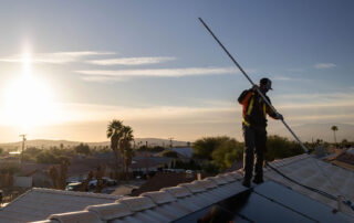 Renovian cleaning solar panels on top of roof, the sun shining in the background and the renova worker is wearing work gear and a long cleaning tool