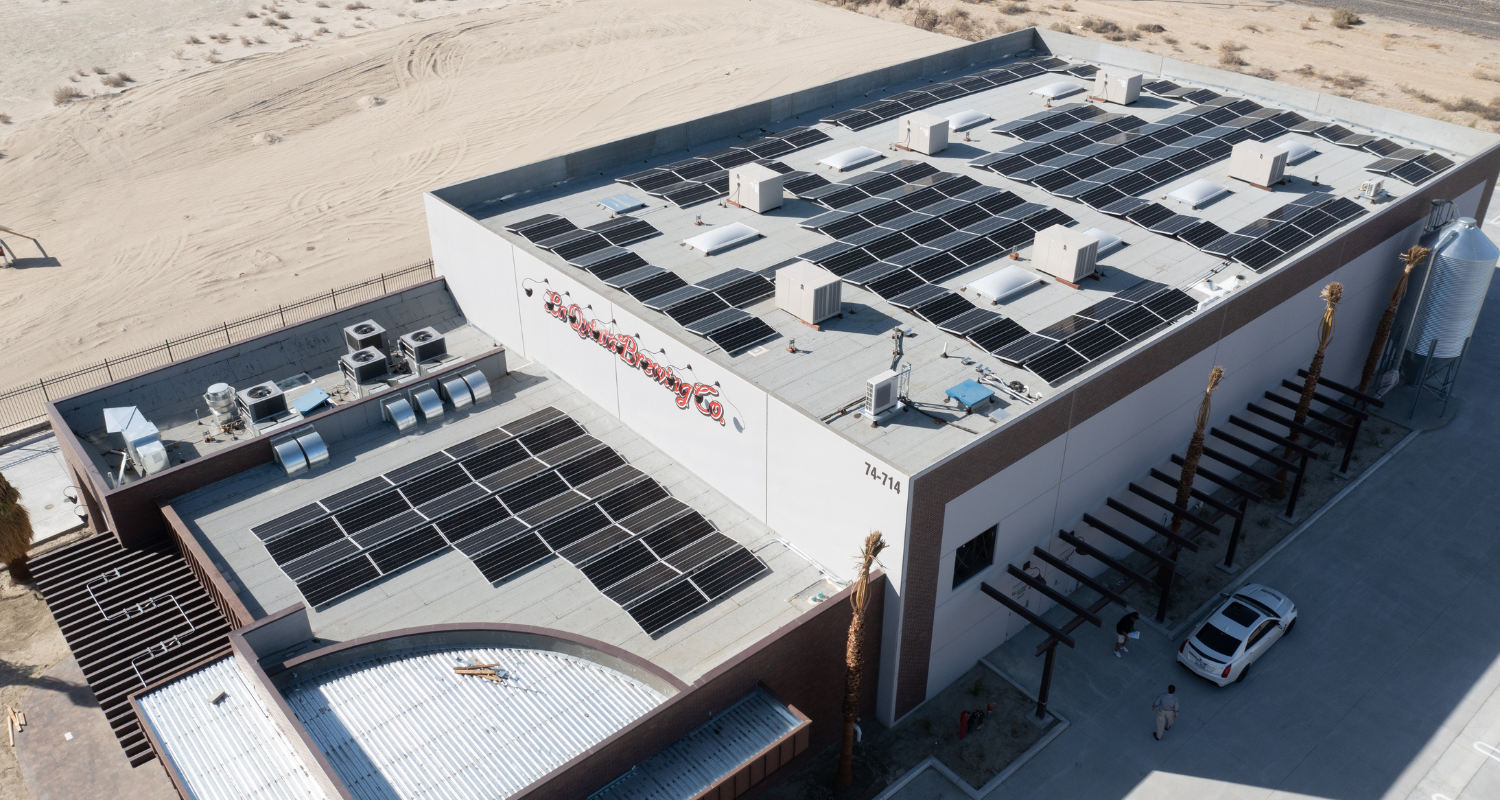 Aerial view of a large industrial building with numerous solar panels on the flat rooftop. The structure is surrounded by sandy terrain and a fence. Several air conditioning units are visible on the roof. A few vehicles are parked near the entrance. The sky is clear.