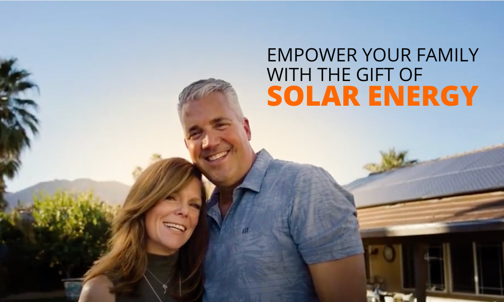 A smiling couple embraces in front of a house with solar panels on the roof. The man has gray hair, and the woman has light brown hair. They stand outdoors with trees and mountains visible in the background. Text reads, Empower your family with the gift of solar energy.