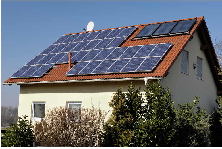 A house with a red-tiled roof features multiple solar panels and a solar water heater. The panels are mounted in rows, covering much of the roof. The building is surrounded by various bushes and trees, set against a clear blue sky. The walls of the house are cream-colored.