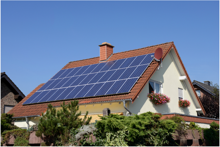 A house with red roof tiles features a large array of solar panels. The building is surrounded by green shrubs and flowers, with two hanging flower baskets on the right. A clear blue sky is in the background.