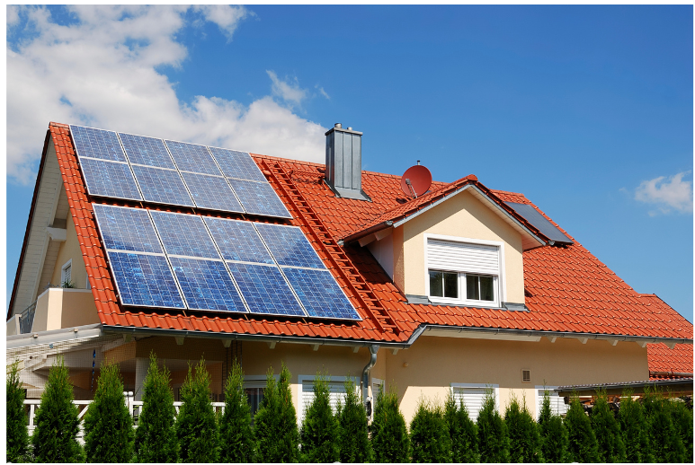 A modern house with a red-tiled roof features multiple solar panels on top. The building is light-colored, with a chimney and a satellite dish. A row of green bushes is in the foreground, and the sky is clear and blue with a few white clouds in the background.