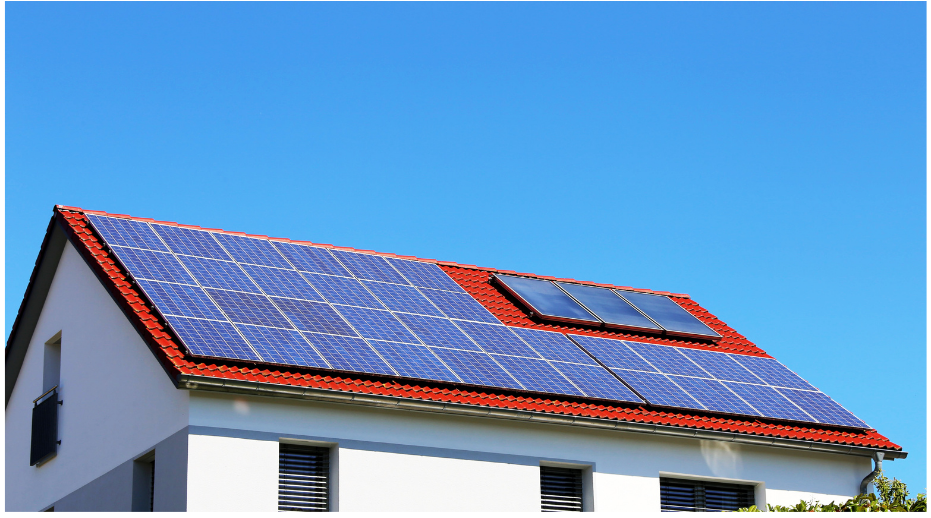 A white house with a red-tiled roof features an array of solar panels installed on top. The panels cover most of the roof, with a clear blue sky in the background, indicating a sunny day. The building has three small windows visible on the side.