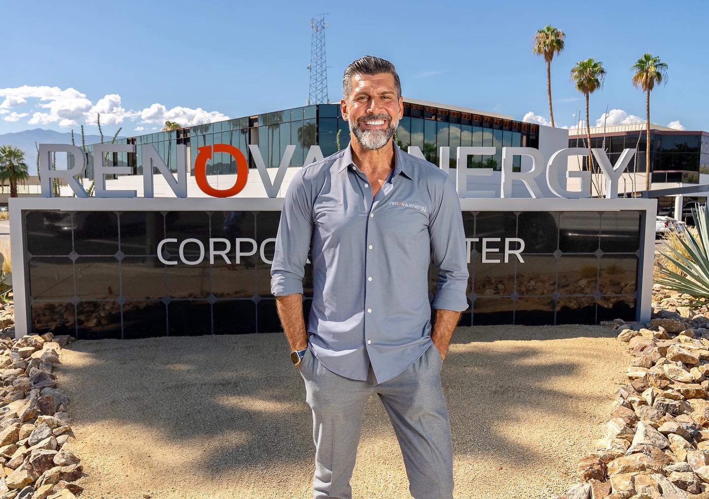 A smiling man in a gray shirt stands outdoors in front of a sign that reads RENOVA ENERGY CORPORATE CENTER. The background features a modern building with large glass windows, palm trees, and a clear blue sky. Rocks and desert landscaping are visible in the foreground.