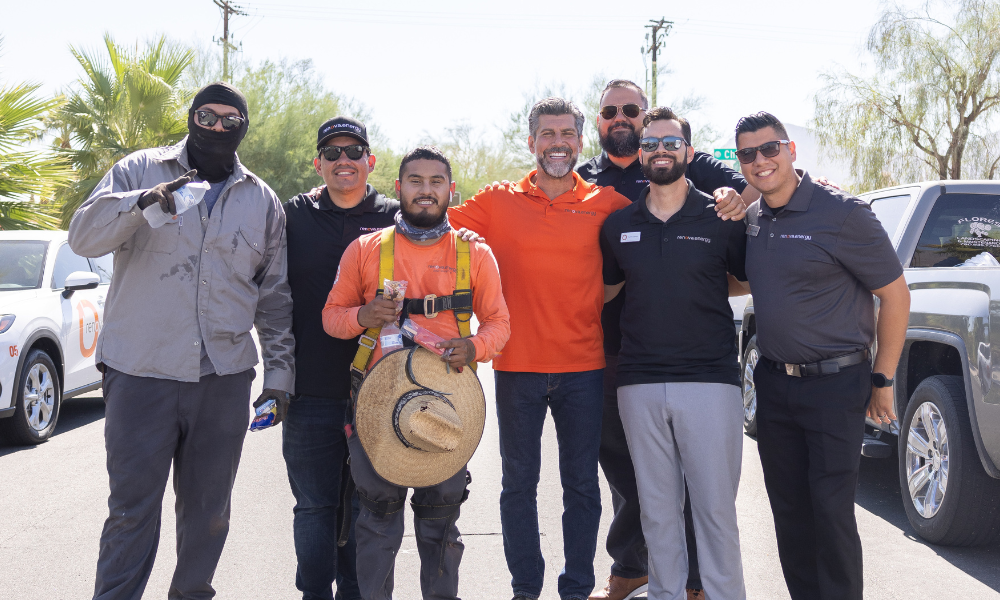 A group of seven men stands together in a sunny outdoor setting, smiling for the camera. They are casually dressed, with two men in safety vests and hard hats. Palm trees and parked vehicles are visible in the background.