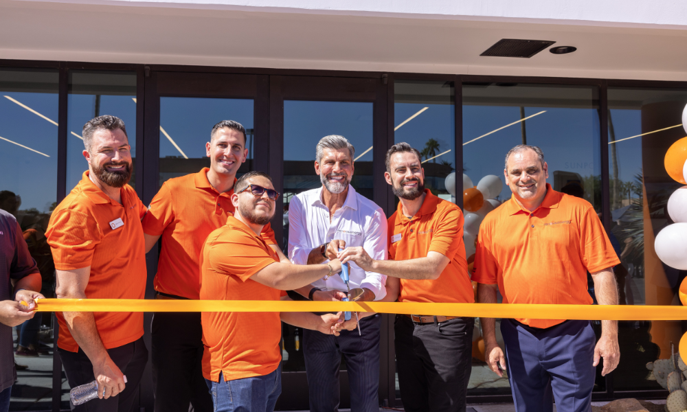 A group of six men stand in front of a buildings entrance during a ribbon-cutting ceremony. Five are wearing bright orange shirts, and the man in the center wears a white shirt. Two hold scissors poised to cut the orange ribbon. White and orange balloons are visible on the side.