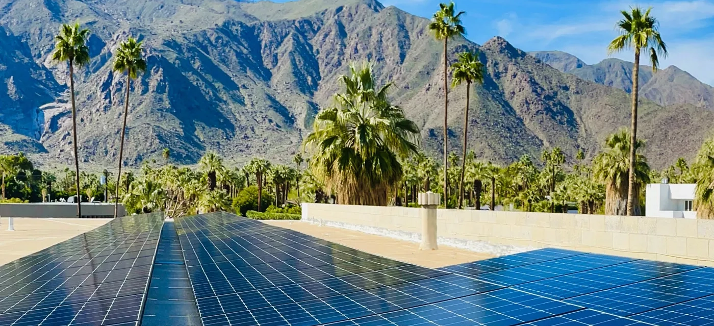 A rooftop covered with solar panels beneath a clear blue sky. In the background, tall palm trees stand against a backdrop of rugged, sunlit mountains. The scene reflects a mix of sustainable technology and natural beauty.