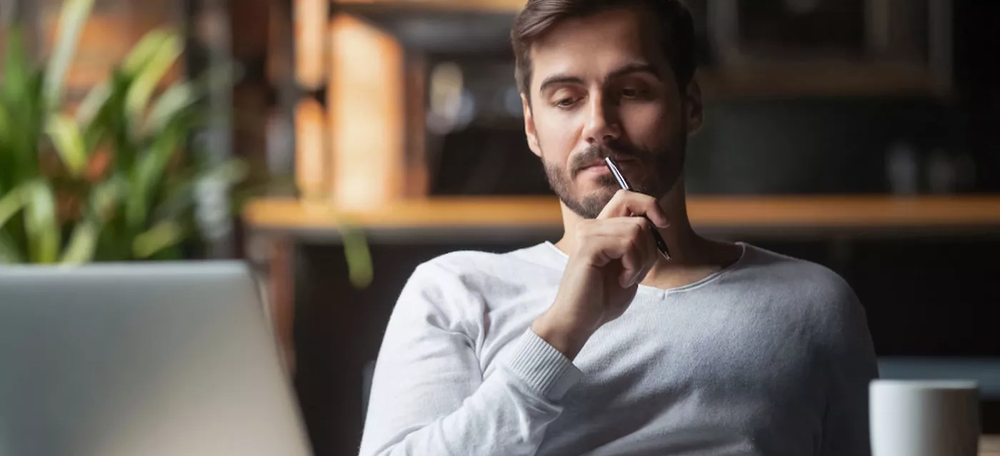 A man sits thoughtfully with a pen to his lips, gazing at a laptop screen. He has short hair and a beard, wearing a light gray sweater. The background is softly blurred, with a plant visible. The setting appears to be a cozy, well-lit indoor space.