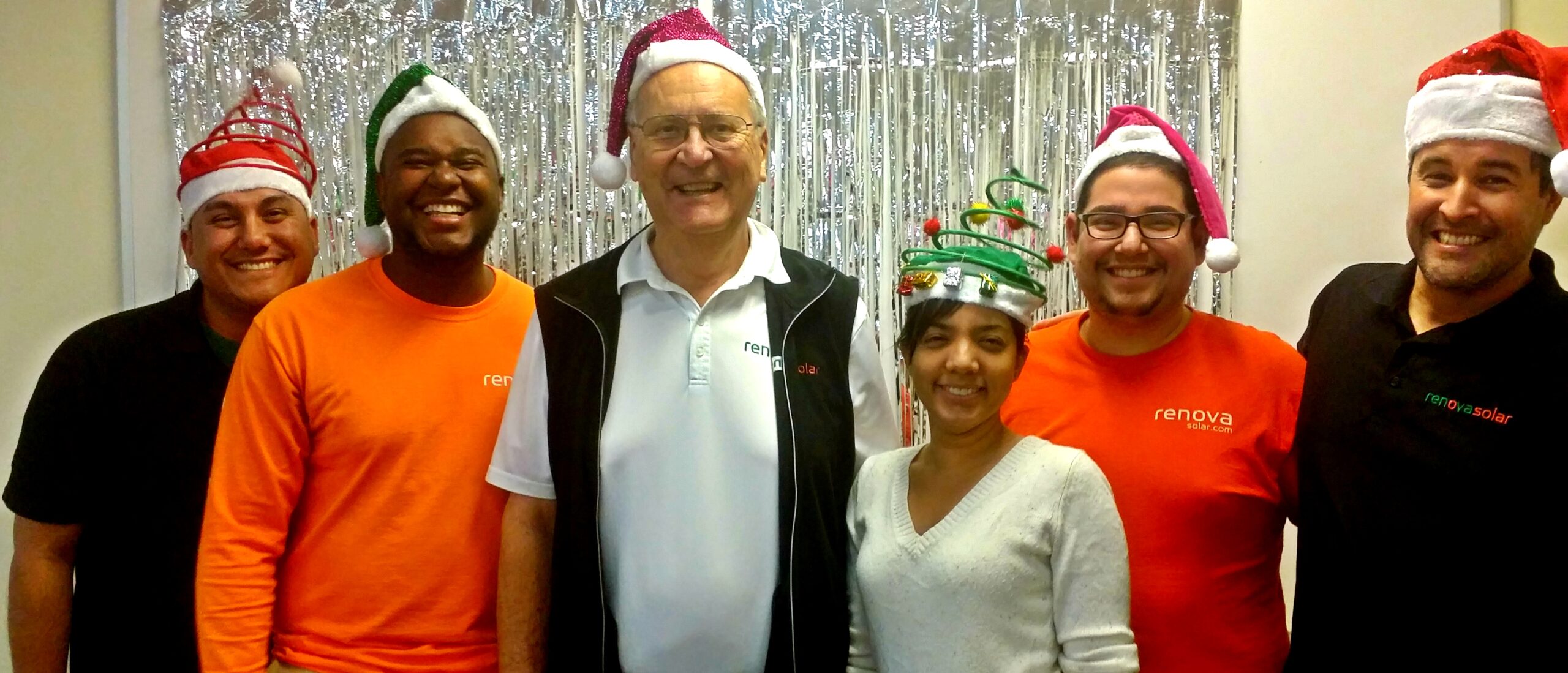 A group of six people stands in a line, all wearing festive Santa hats. They are smiling, with some wearing renova branded shirts. The background features silver tinsel decorations, suggesting a holiday celebration.