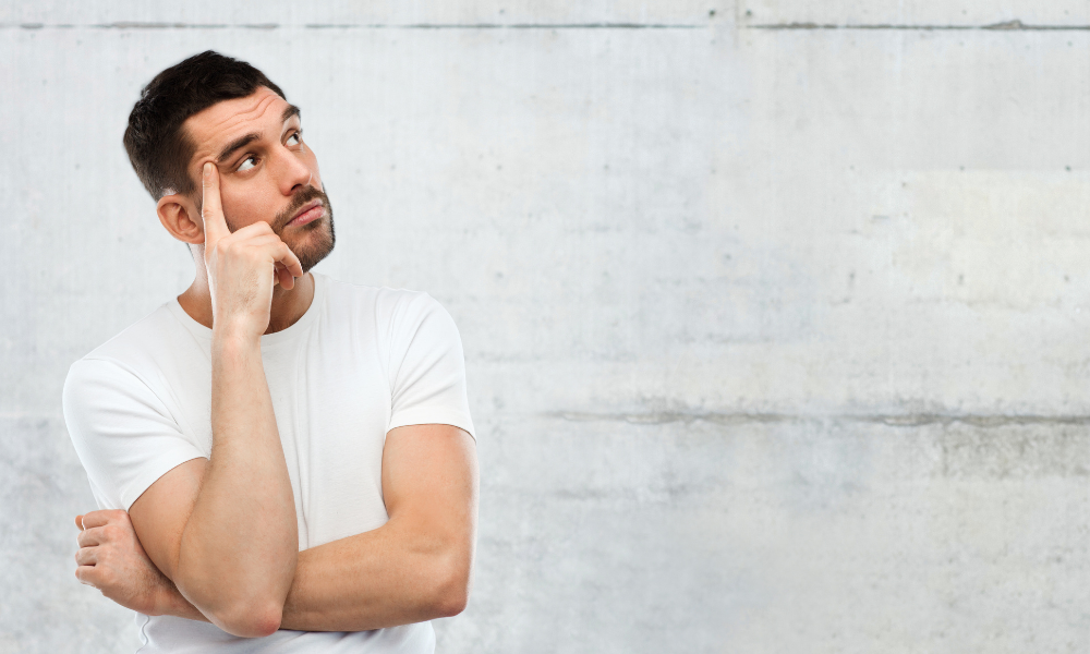 A man in a white t-shirt stands against a light gray concrete wall, with a thoughtful expression. He gazes upward, touching his temple with his right index finger, and his left arm is crossed over his chest.