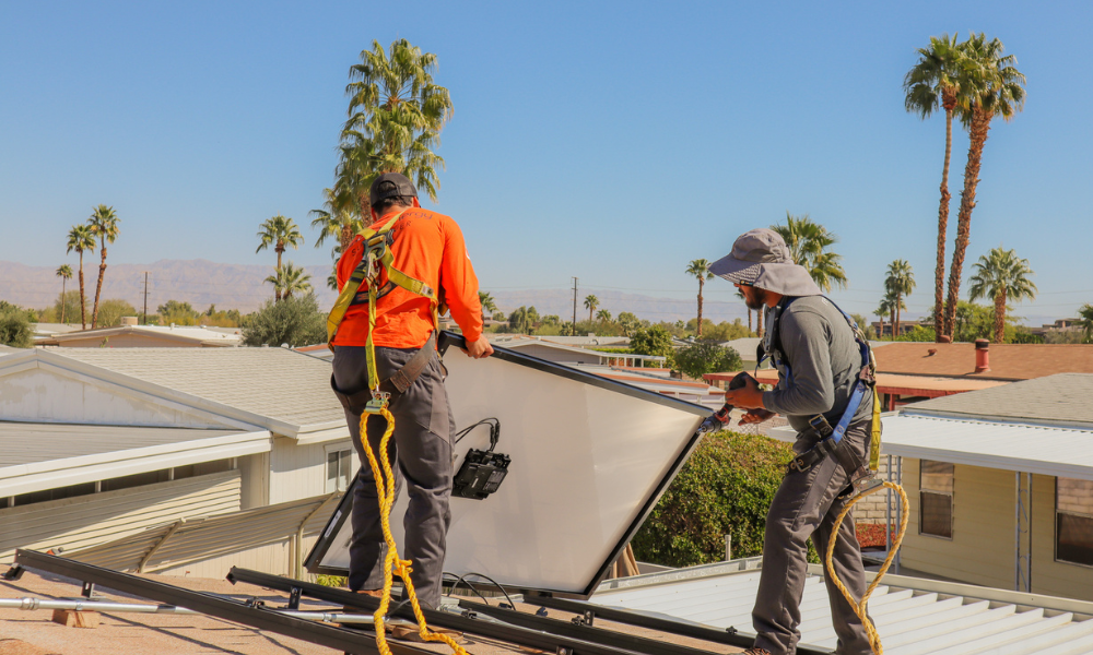 Two workers install solar panels on a rooftop under a clear blue sky. Both wear safety harnesses and sun hats. Palm trees and rooftops are visible in the background, along with distant mountains. The worker on the left wears an orange shirt, while the one on the right wears a gray shirt.