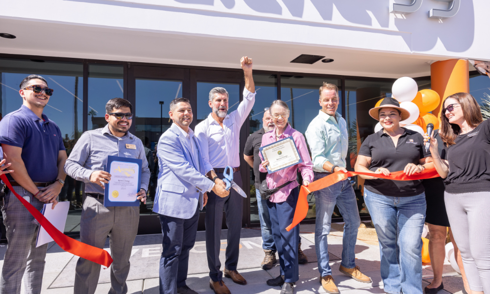 A group of people stand in front of a building for a ribbon-cutting ceremony. Two men hold certificates, another raises his arm in celebration. A red ribbon is stretched across, and two women prepare to cut it. Balloons in white and orange are on display, with a sunny backdrop.