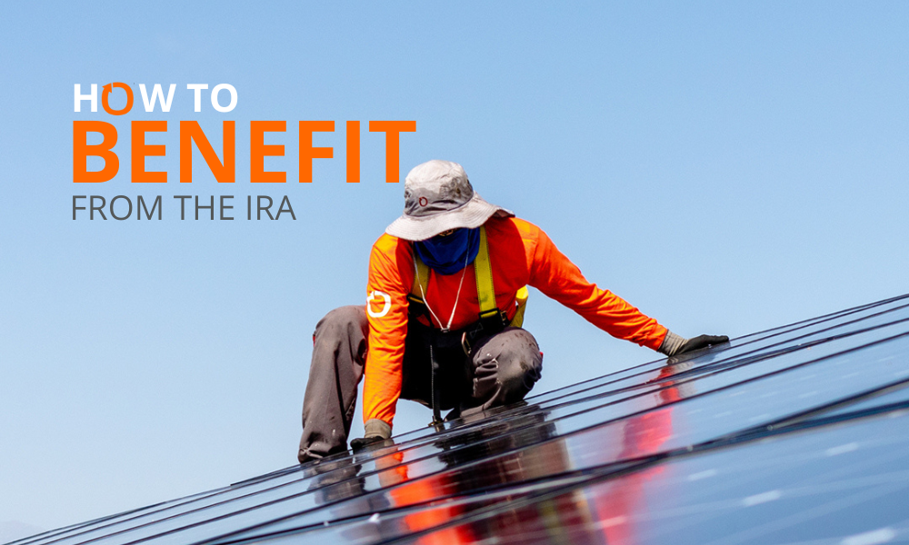 A worker in an orange long-sleeve shirt and wide-brim hat is installing solar panels on a rooftop under a clear blue sky. Bold text reads How to Benefit from the IRA in the upper left corner. The scene highlights renewable energy and incentives.