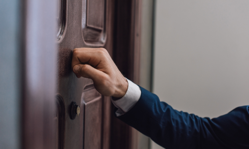 A person wearing a blue suit is knocking on a wooden door with their right hand. The door is brown with rectangular panels, and a peephole is visible near the top left. The scene suggests a professional or formal visit.