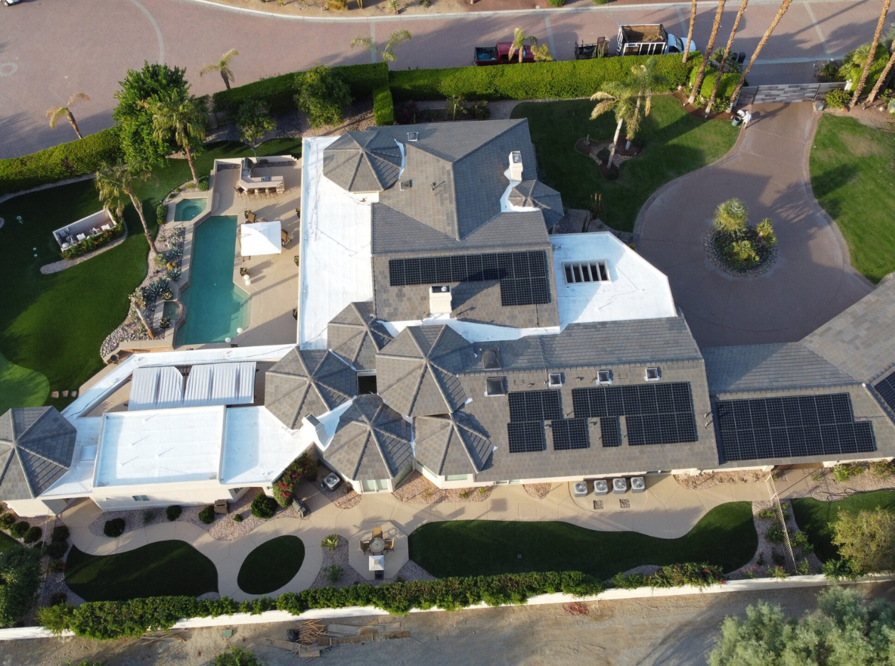 Aerial view of a large house with solar panels on the roof. The property features a swimming pool with a shaded area, surrounded by landscaped greenery and palm trees. A circular driveway provides access to the house, with well-maintained garden paths and decorative elements visible.