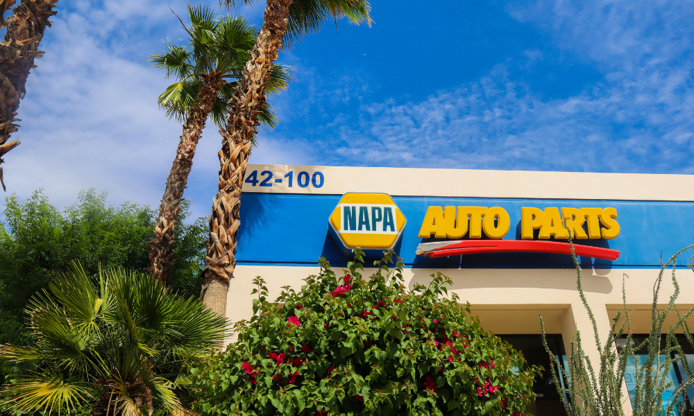 A NAPA Auto Parts store facade with its blue and yellow logo and sign against a blue sky. Palm trees and lush greenery surround the building, which is painted in cream and blue tones. The stores address, 42-100, is displayed above the entrance.