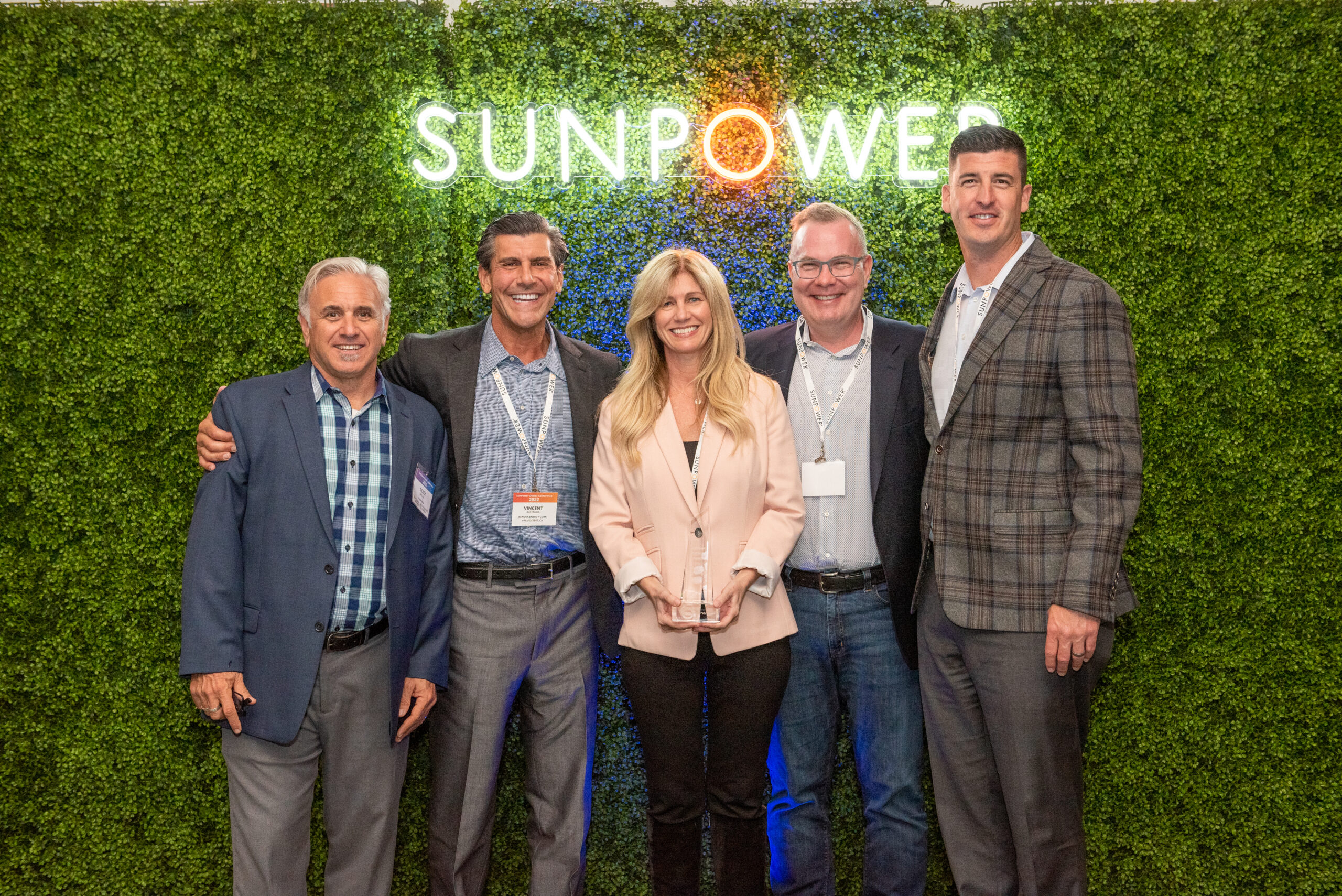 Five people stand smiling in front of a green foliage wall featuring the illuminated word SUNPOWER. They are dressed in business attire. The person in the middle holds a plaque or award.