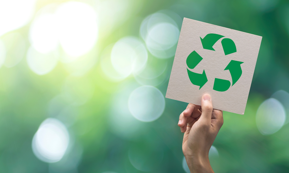 A hand holds a square piece of paper with the green recycling symbol on it. The background features a blurred, lush, green bokeh effect, suggesting a natural environment and emphasizing the theme of sustainability.