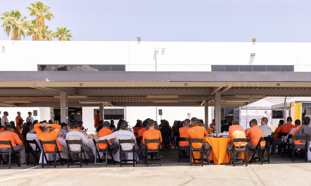 A group of people wearing orange shirts sit under a shaded outdoor area for a meeting. They face a speaker outside the frame. The building behind them is white with rectangular windows. A few palm trees are visible under a clear blue sky. Tables with orange cloths are scattered around.