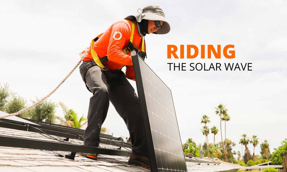 A worker in an orange safety vest and wide-brimmed hat installs a solar panel on a rooftop. They are secured with a harness. The sky is clear, and palm trees are visible in the background. The text Riding the Solar Wave is displayed to the right of the worker.