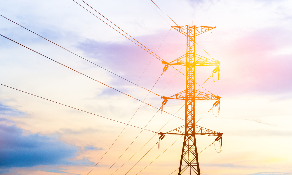 A tall electricity transmission tower stands against a vibrant sky during sunset. The sky features a gradient of colors ranging from blue to orange and pink, with streaks of clouds. The metallic structure of the tower is silhouetted, and multiple electrical wires extend from it.