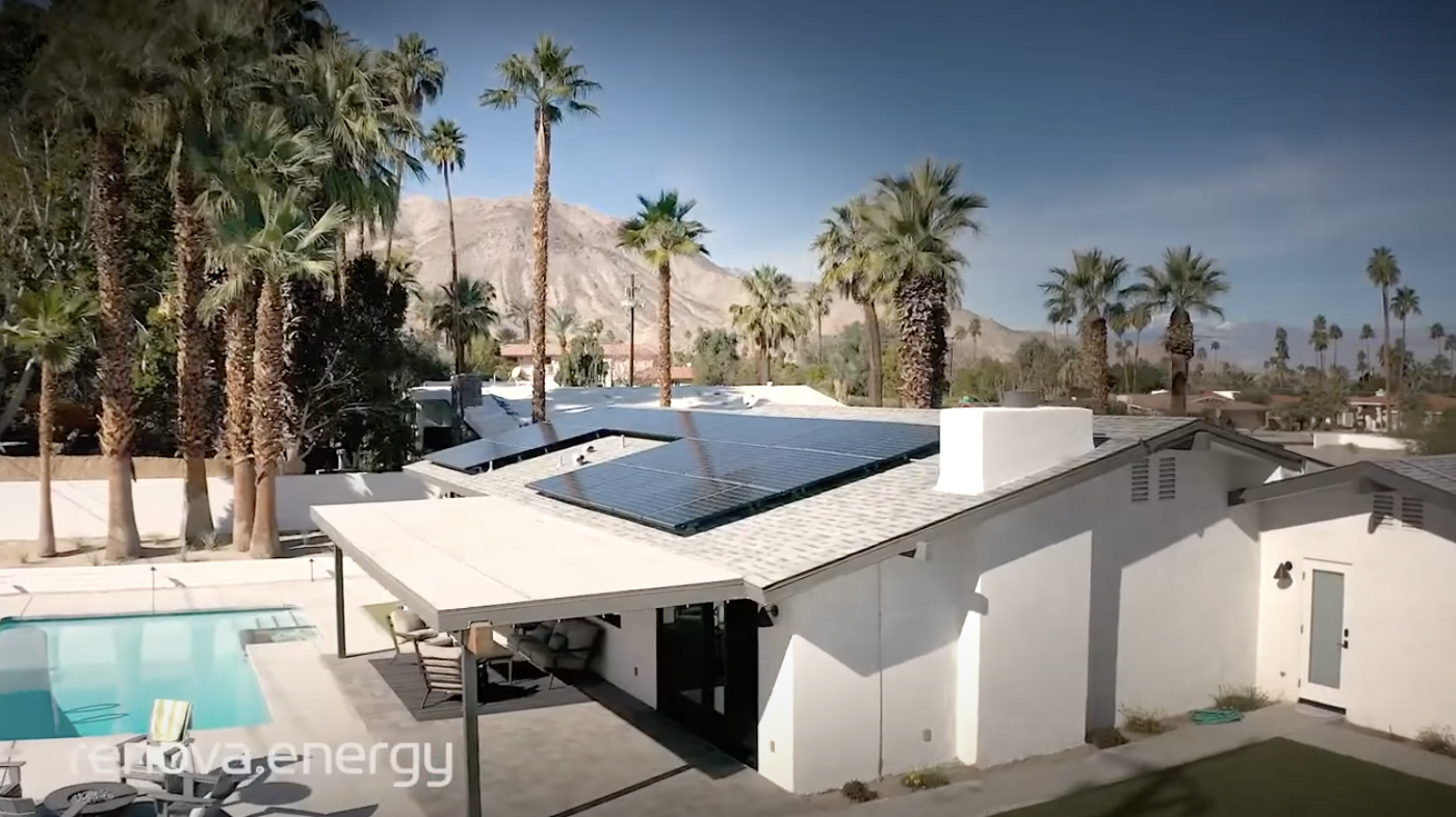 A modern white house with solar panels on the roof is surrounded by tall palm trees and a clear blue sky. A swimming pool is visible in the backyard, along with patio furniture. Mountains can be seen in the background, creating a serene and sunny landscape.