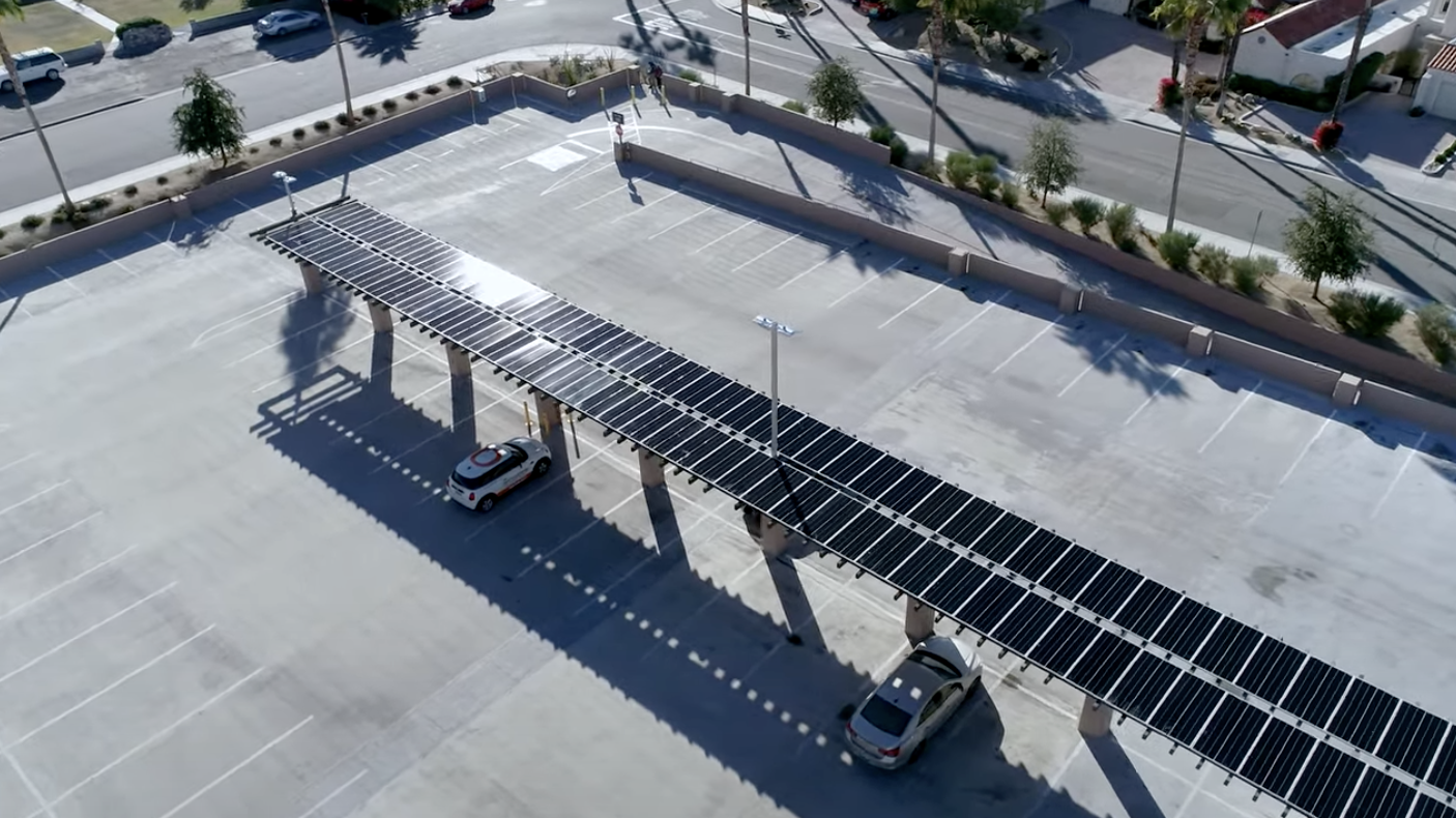 Aerial view of a parking lot with an array of solar panels mounted on a long canopy. Two cars are parked underneath, benefiting from the shade. Surrounding the lot are roads and palm trees, characteristics of a sunny environment.