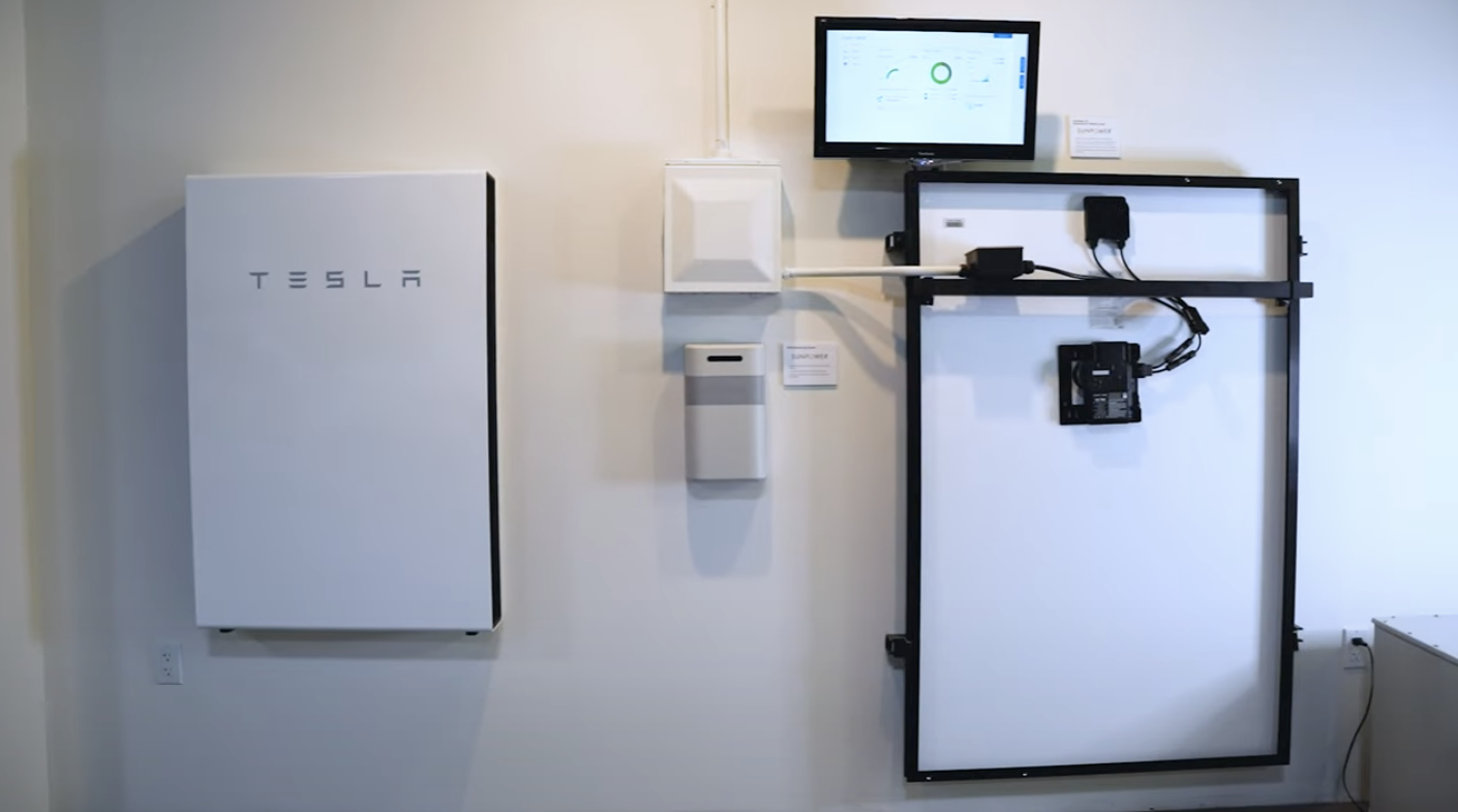 A Tesla Powerwall, a rectangular white device mounted on a wall, is next to electrical components and a screen displaying energy data. The screen and components are organized neatly, with the setup showcasing a residential energy storage system.