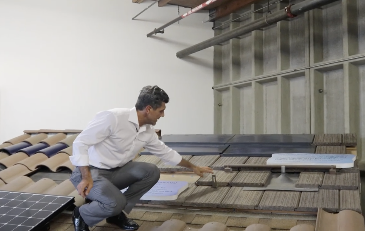 A man in a white shirt and gray pants kneels on a display roof, examining different types of roof tiles and solar panels. The background shows a white wall and metal pipes overhead.