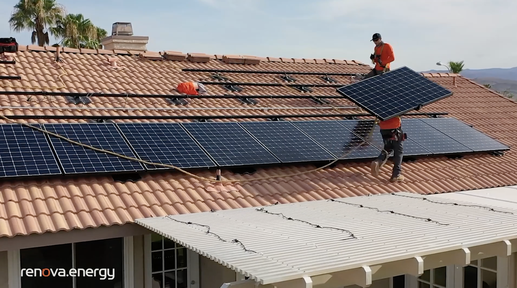 Workers in orange shirts install solar panels on a terracotta-tiled roof of a residential home. One worker positions a large solar panel, while another adjusts mounting hardware. The roof has a white pergola at the edge. Hills and a clear sky are in the background.