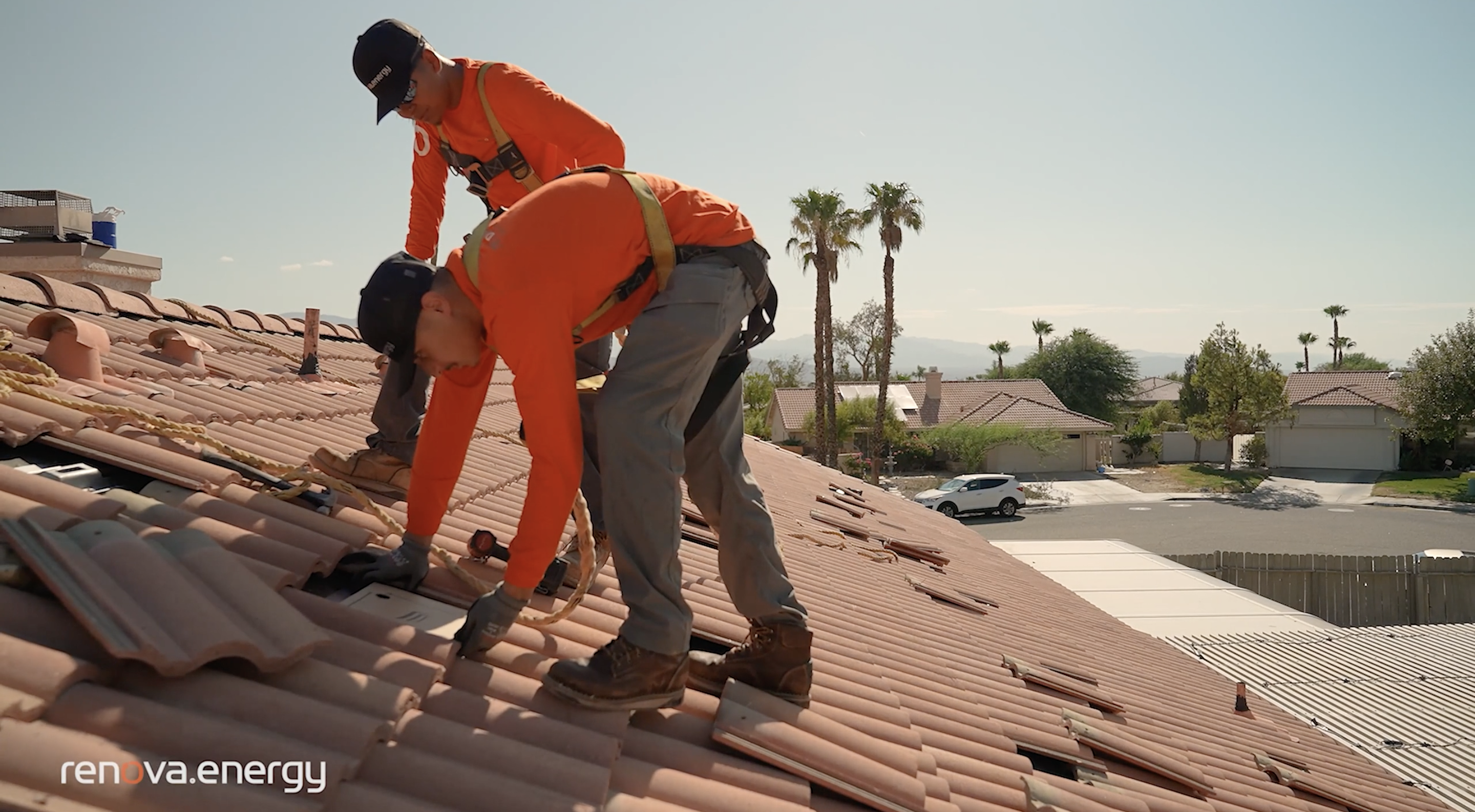 Two workers in orange shirts and safety harnesses install solar panels on a tiled roof under a clear sky. Houses, palm trees, and mountains are visible in the background. A white van is parked on the street below. The workers are focused and equipped with tools.