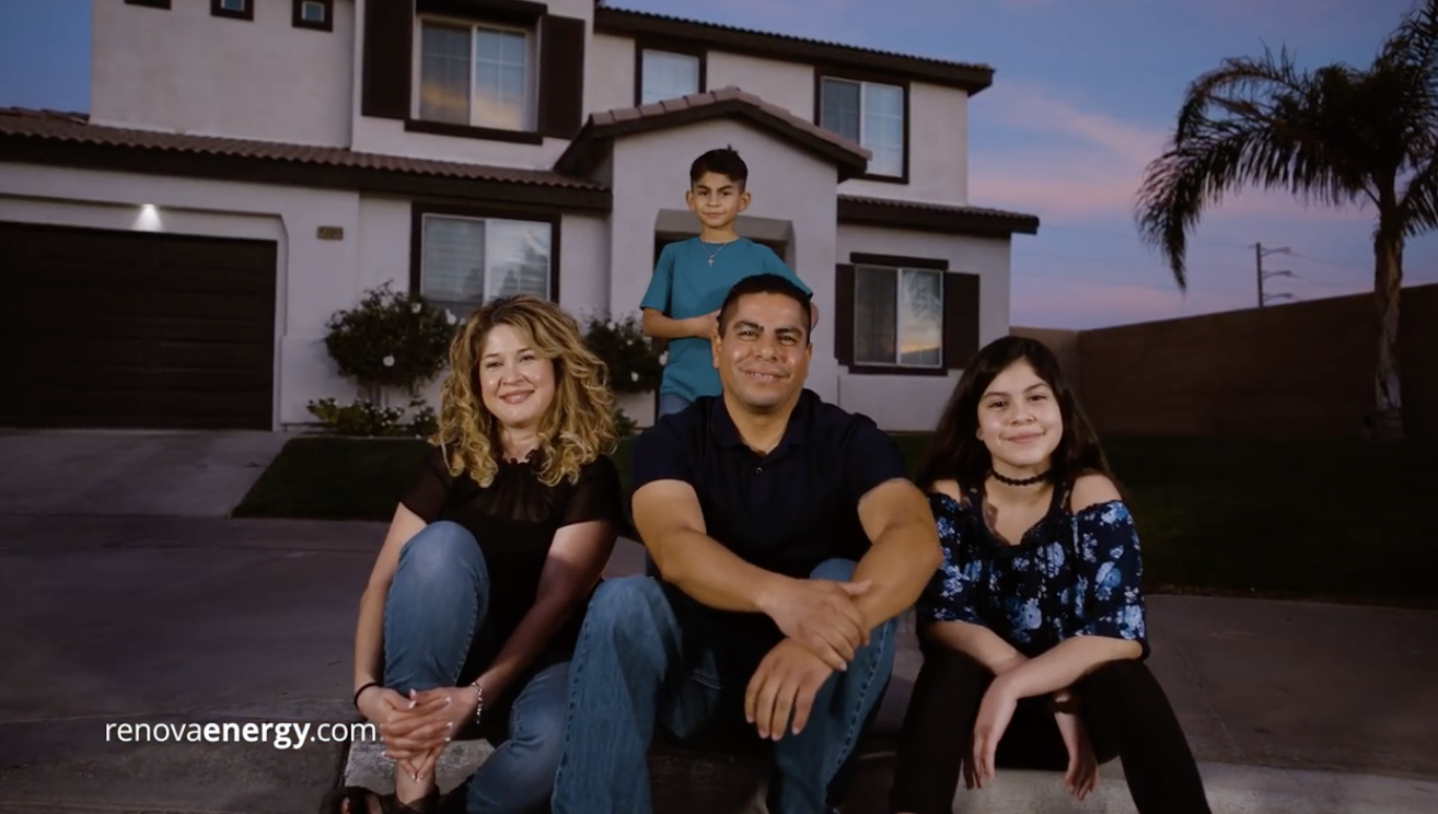 A family of four sits on the curb in front of a two-story house at dusk. The parents and daughter smile at the camera while the son stands behind them. The adults wear jeans and dark tops; the daughter wears a blue floral blouse, and the son is in a green shirt. The text reads renovaenergy.com.