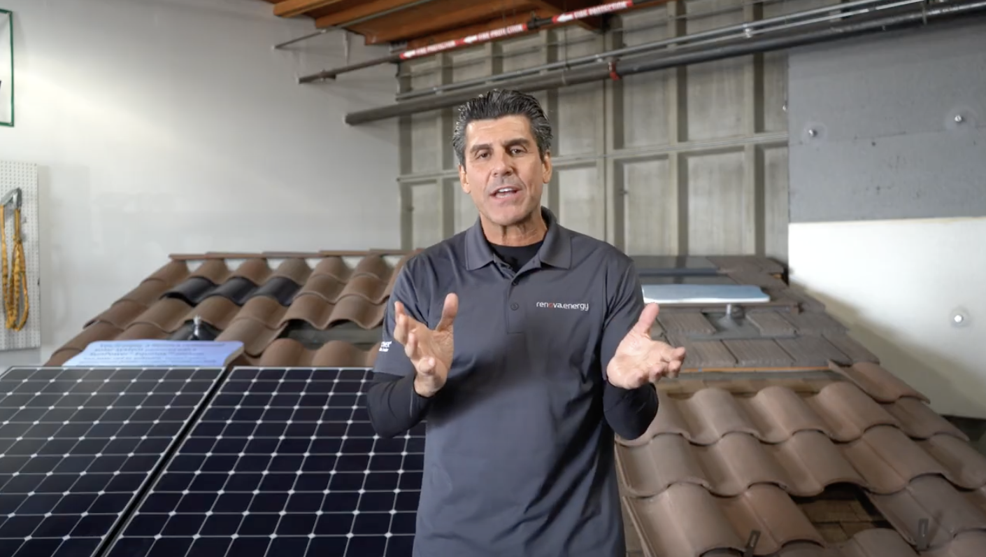 A man gestures while speaking, standing indoors in front of a demonstration setup featuring solar panels and roof tiles. Hes wearing a gray polo shirt with red lettering and has short, dark hair. The background includes a tiled roof model, panels, and metal piping overhead.