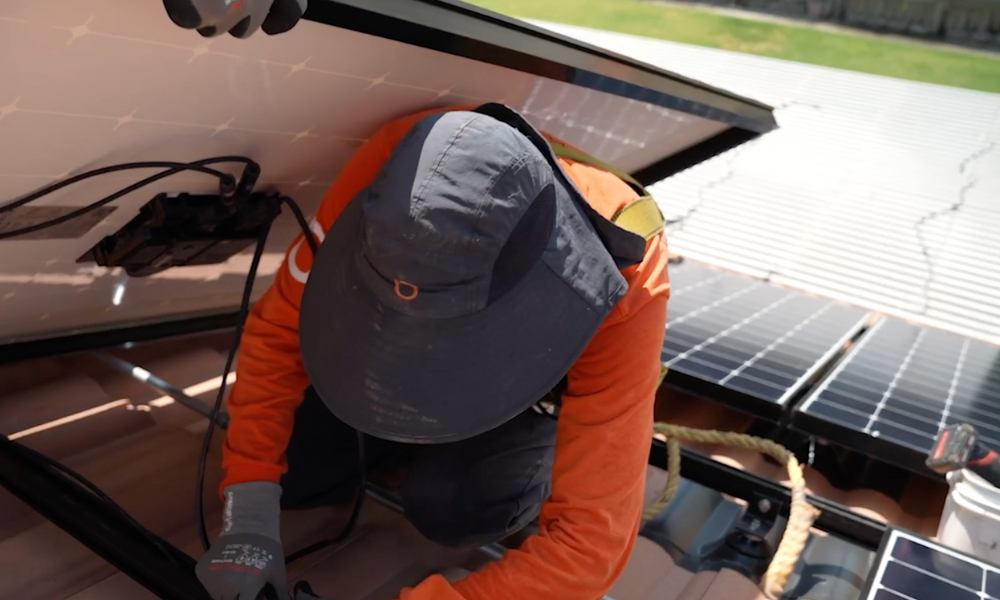 A person in an orange long-sleeve shirt and wide-brimmed hat installs solar panels on a rooftop. They are kneeling and working with electrical components under a tilted solar panel. The background shows more panels and part of the roof. Safety equipment and ropes are visible beside them.