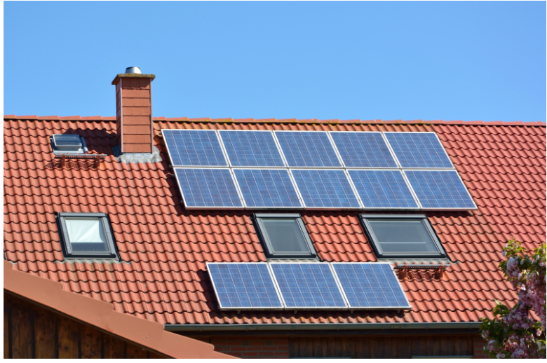 A red-tiled roof with several solar panels mounted on top. There is a brick chimney on the left side, two skylights near the center, and a clear blue sky in the background. A small portion of green foliage is visible on the bottom right.