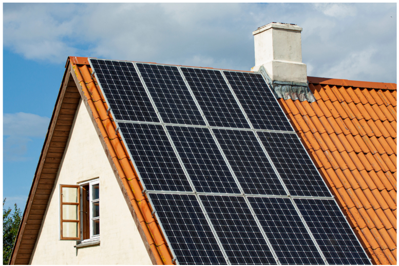 A house with a sloped red-tiled roof features multiple solar panels arranged in rows. A chimney is visible on the roofs peak. An open window is on the side of the house, and the sky is partly cloudy, indicating a sunny day.