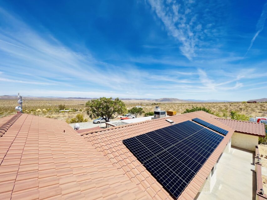 A house with a red-tiled roof features solar panels on one section. The surrounding landscape is a vast, arid desert under a clear blue sky with wispy clouds. A few scattered trees and parked vehicles are visible near the house, extending into the expansive, dry terrain.