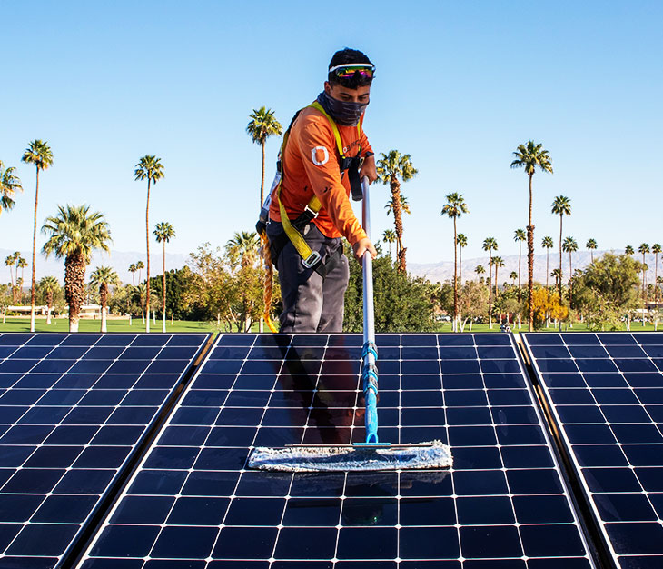 A worker is cleaning solar panels with a long-handled brush under a clear blue sky. The worker is wearing an orange long-sleeve shirt, cap, gloves, and face covering. Palm trees are visible in the background, suggesting a sunny location.