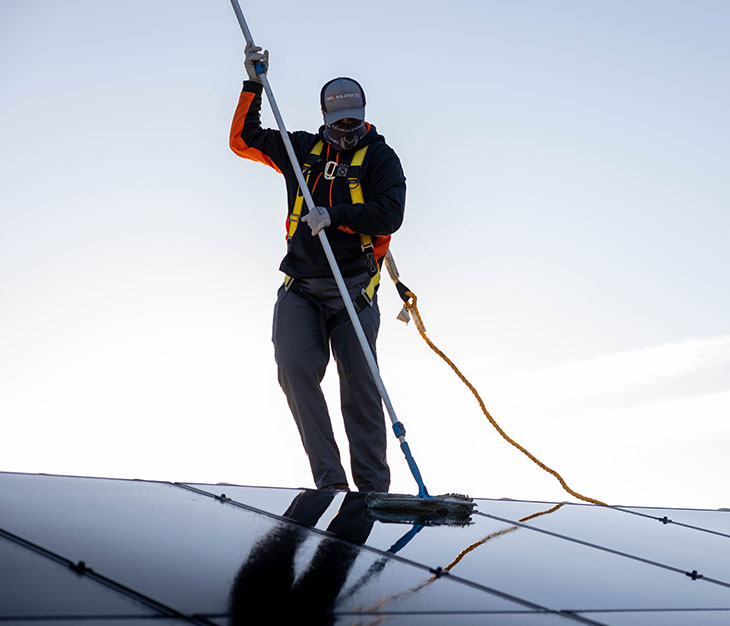 A person wearing safety gear and a harness cleans solar panels with a long-handled brush. The individual is on a rooftop against a clear sky, with the sun casting a bright light. They are dressed in dark clothing and a cap, emphasizing safety and maintenance of renewable energy sources.