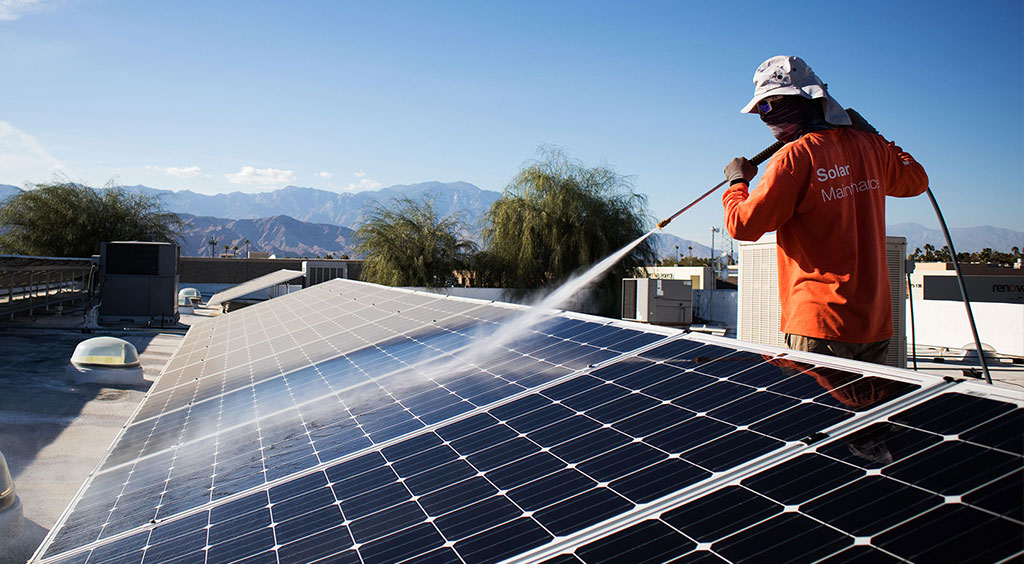 A worker in an orange shirt is cleaning solar panels on a rooftop under a clear blue sky. The person is using a water hose while wearing a hat and face covering. In the background, there are mountains and trees. The shirt has a logo and text, but details are not legible.