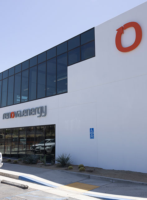 A modern, two-story building with large glass windows and a logo featuring an orange circle on the upper right corner. The name Renova Energy is displayed on the white facade. Theres a parking area with a handicap sign, and small desert plants line the walkway.