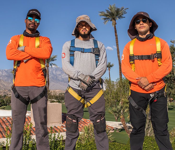 Three men stand confidently in safety harnesses and gear, ready for outdoor work. They wear hats and sunglasses for sun protection. The background features palm trees, a clear blue sky, and a glimpse of a tiled roof, suggesting a warm climate.