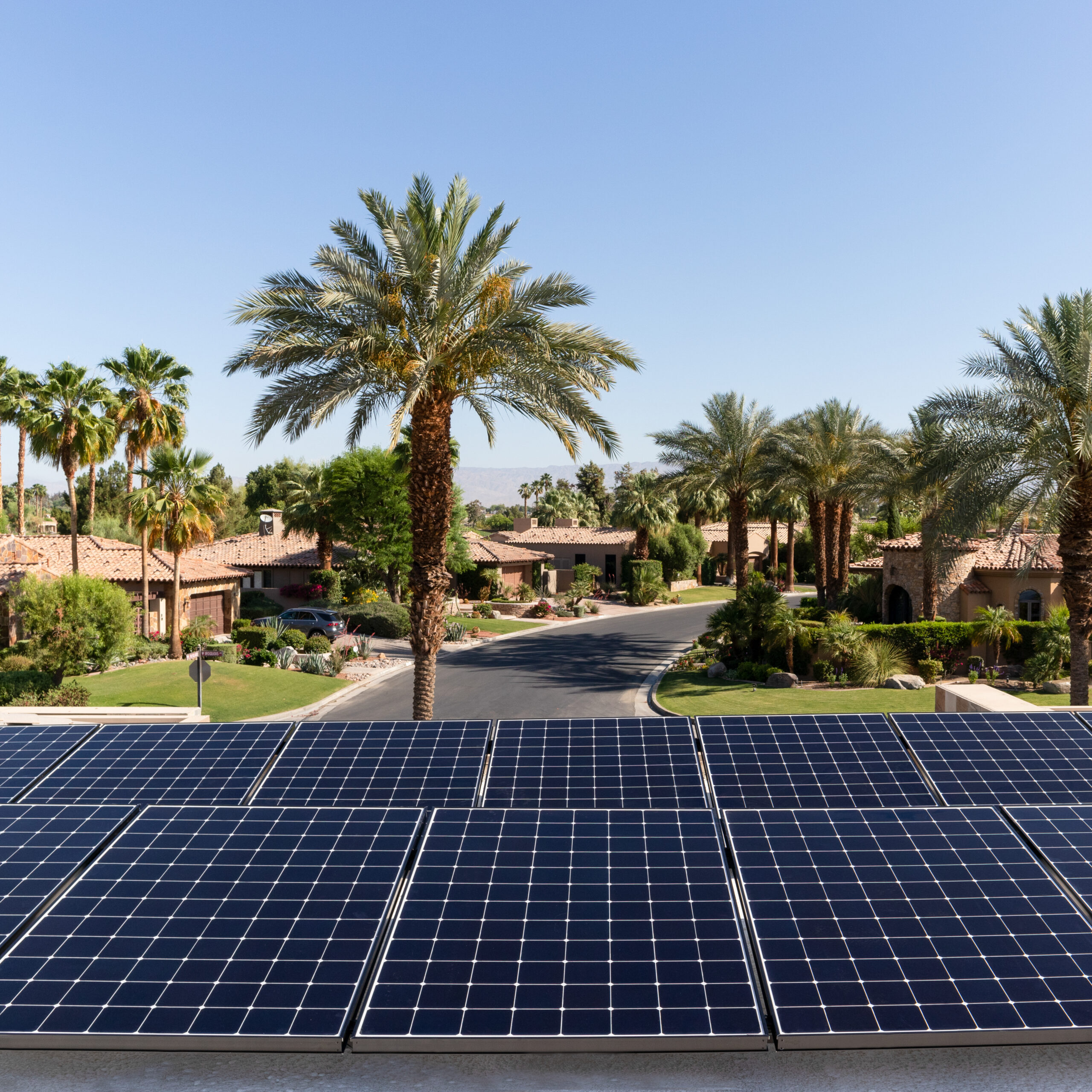 A row of solar panels installed on a rooftop overlooks a suburban neighborhood with palm trees lining the street. The houses have a Mediterranean architectural style, featuring clay tile roofs. The sky is clear and blue, suggesting a sunny day.