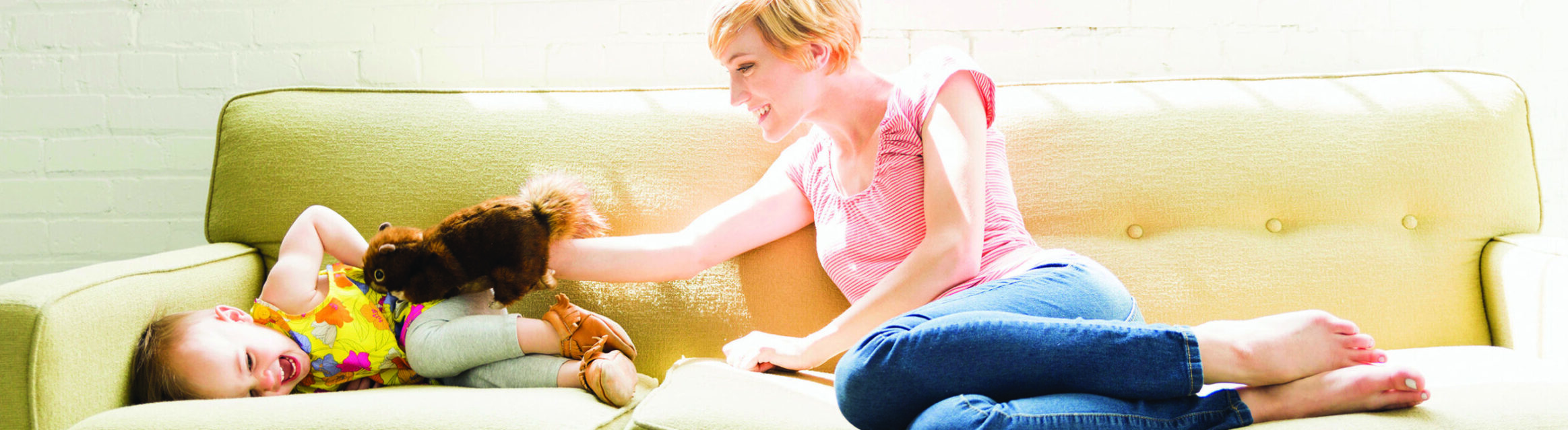 A woman with short hair and a striped shirt playfully holds a toy squirrel towards a laughing child lying on a light green sofa. The child is wearing a yellow and green outfit. Both are enjoying a bright, sunny moment in a cozy room.