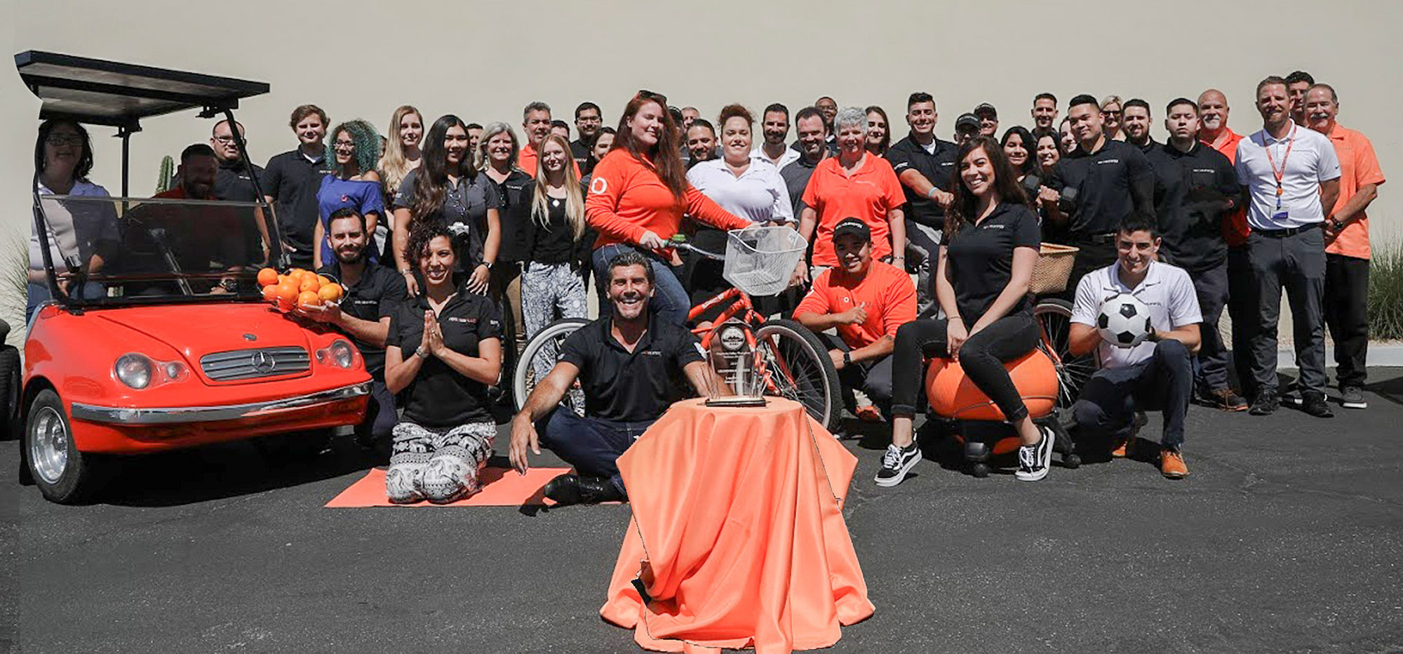 A group of around 40 people poses outside, many wearing orange and black. Some hold basketballs and soccer balls. A red golf cart is on the left. In the center, there is a woven basket and a small trophy on a pedestal. The background is a plain, light-colored wall.