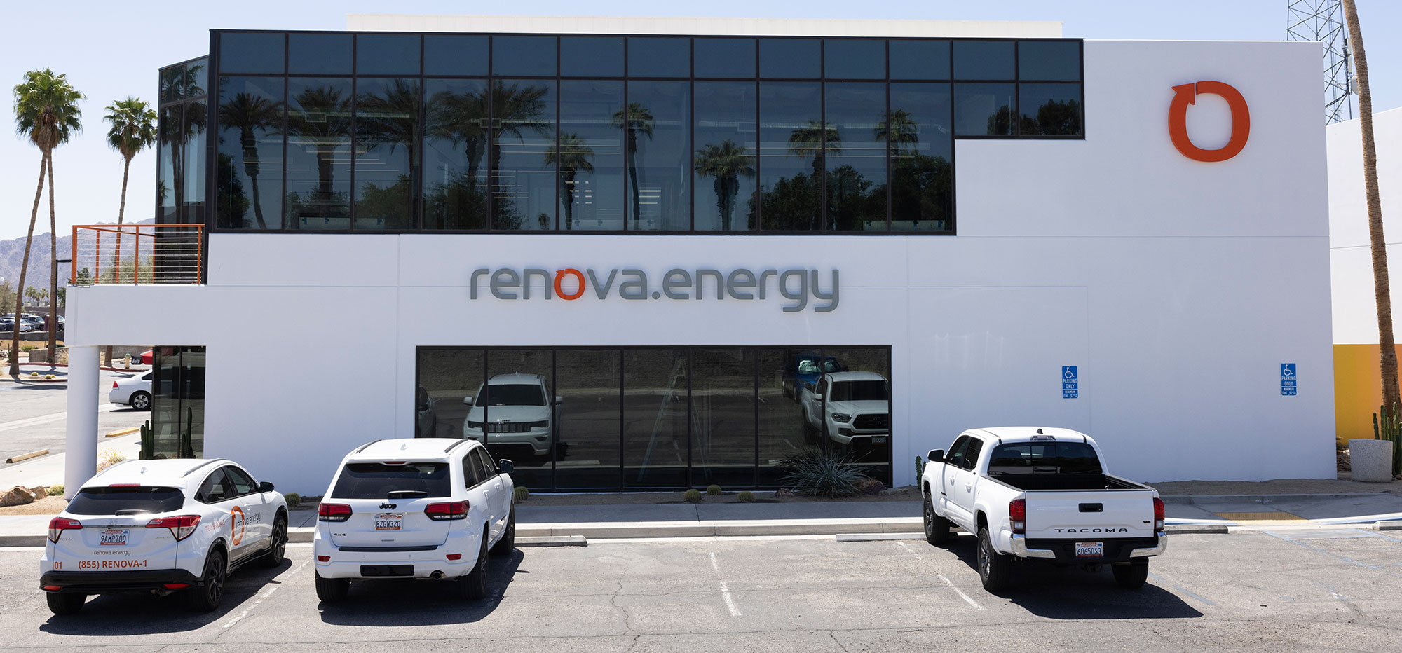 A modern building with a glass facade displays the logo renova.energy in bold letters. Three white vehicles are parked in front of it. Palm trees and a clear sky are visible in the reflection of the windows. The structure has a minimalistic design with a primarily white exterior.