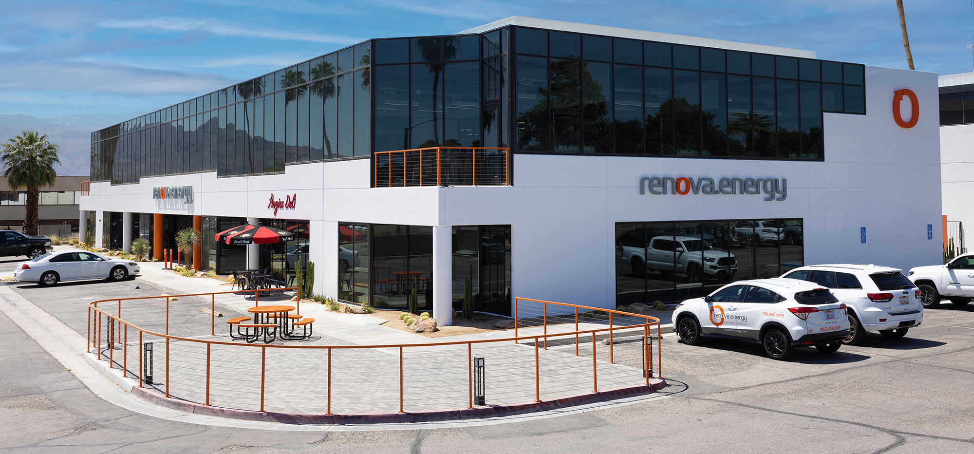 A modern two-story building with large glass windows and Renova Energy signage. Several white company vehicles are parked outside. An outdoor seating area is enclosed by an orange railing, featuring a table with an umbrella. The sky is clear with a few clouds.